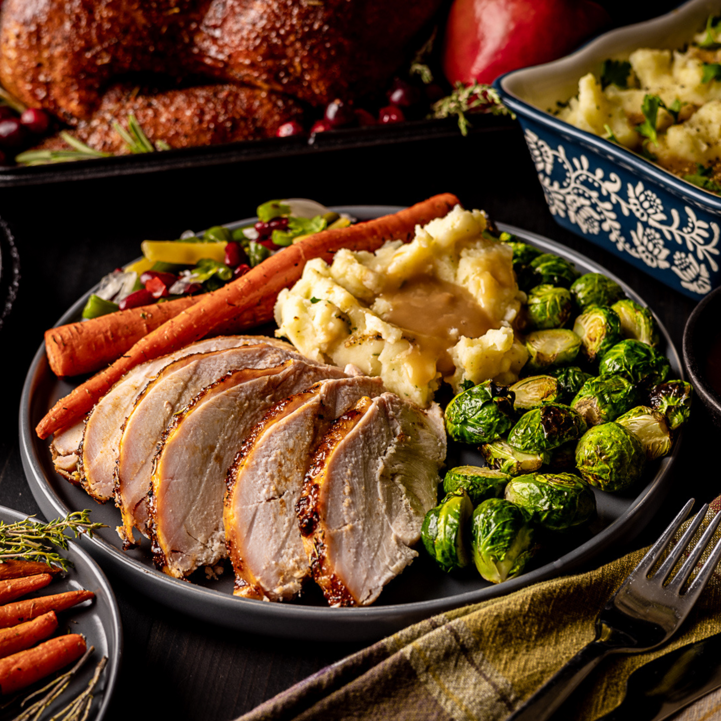 A close up shot of a plate full of food on a dinner table. The plate contains sliced smoked Turkey Breast, grilled Brussels Sprouts, Smoked mashed potatoes and gravy, two carrots, and a green salad.