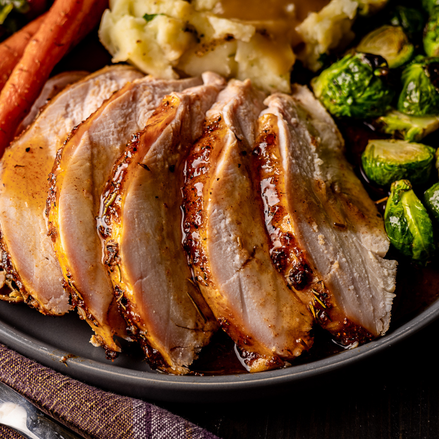 Sliced, roasted turkey breast covered in Cranberry BQQ Glaze sits on a plate with other side dishes including brussels sprouts, and potatoes.  