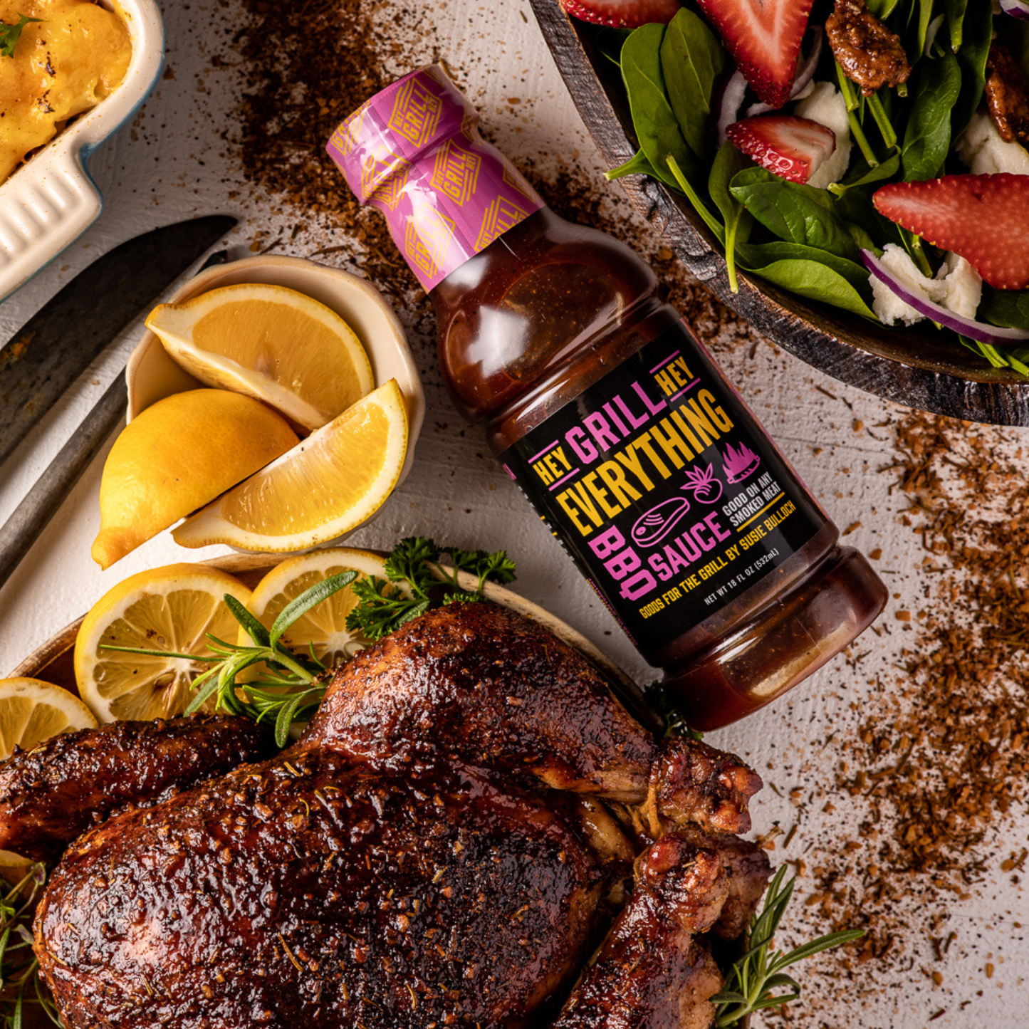 a bottle of Hey Grill Hey Everything Sauce lying flat on a table, surrounded by a whole cooked chicken, a small bowl of sliced lemons, and a large bowl containing a green salad topped with sliced strawberries.