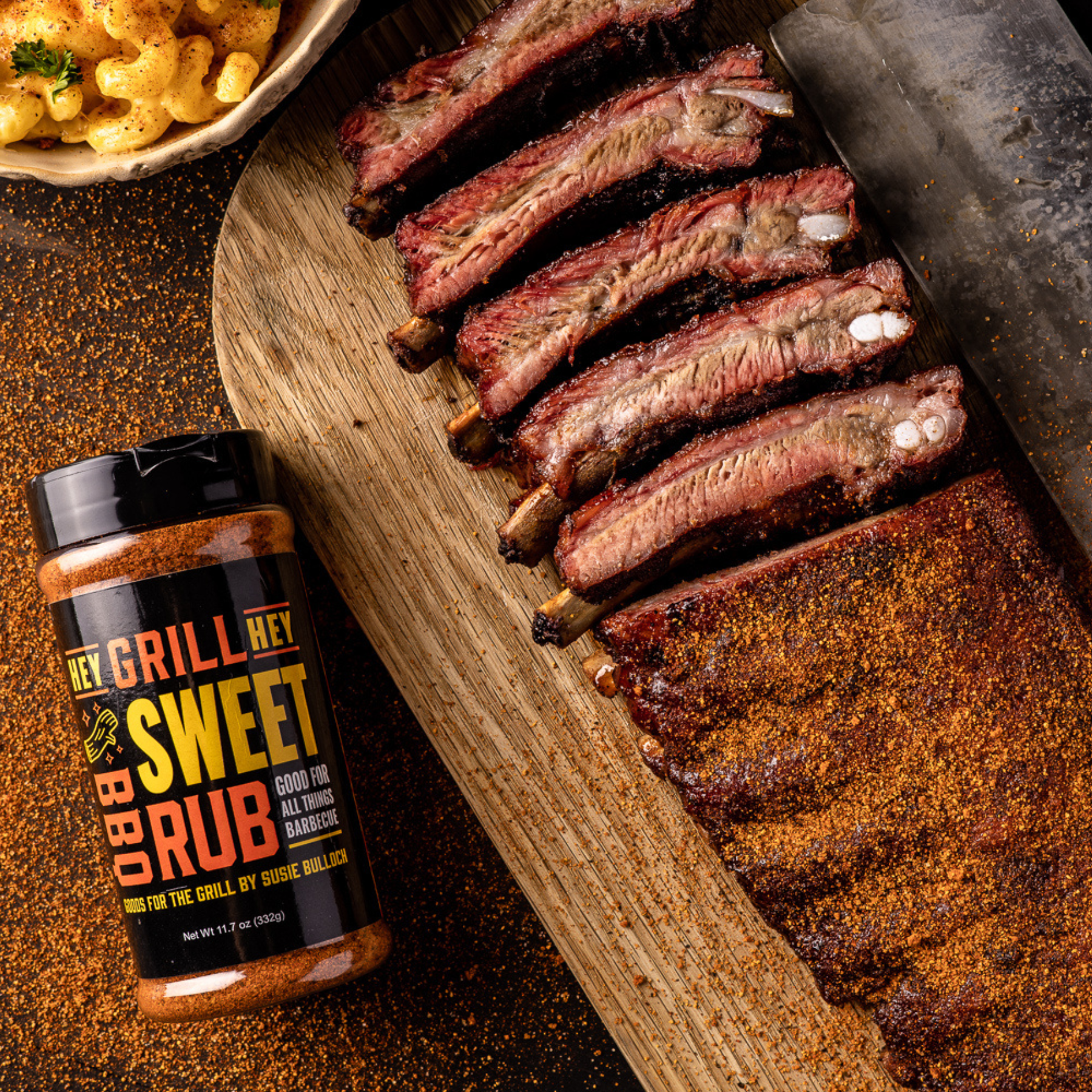 A cooked rack of pork ribs, half of the rack has been sliced, and placed on a wooden cutting board. Next to the board is a bottle of Hey Grill Hey Sweet Rub.
