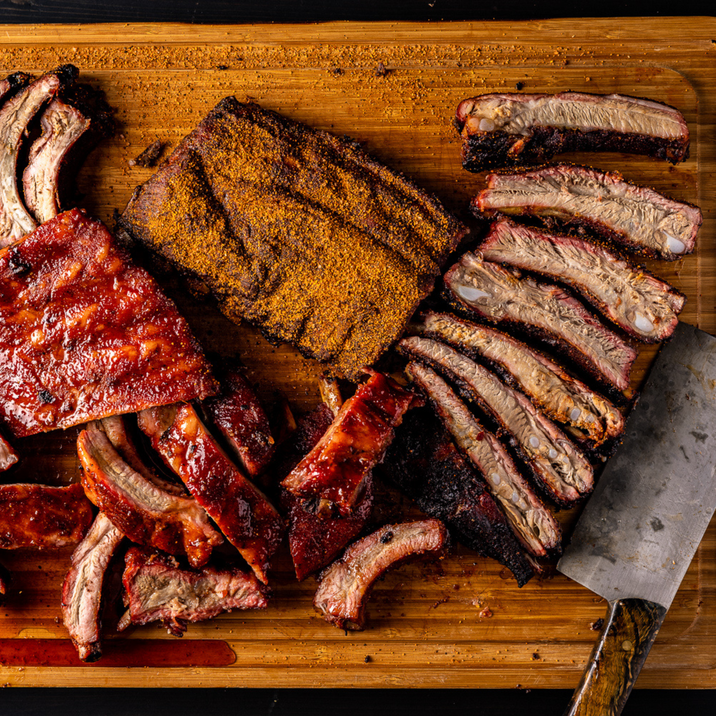 Two different racks of ribs that have been cooked and placed on top of a wooden cutting board with a meat cleaver resting next to it. One rack has been covered in BBQ Sauce, and the other rack of ribs is covered in a dry rub. 
