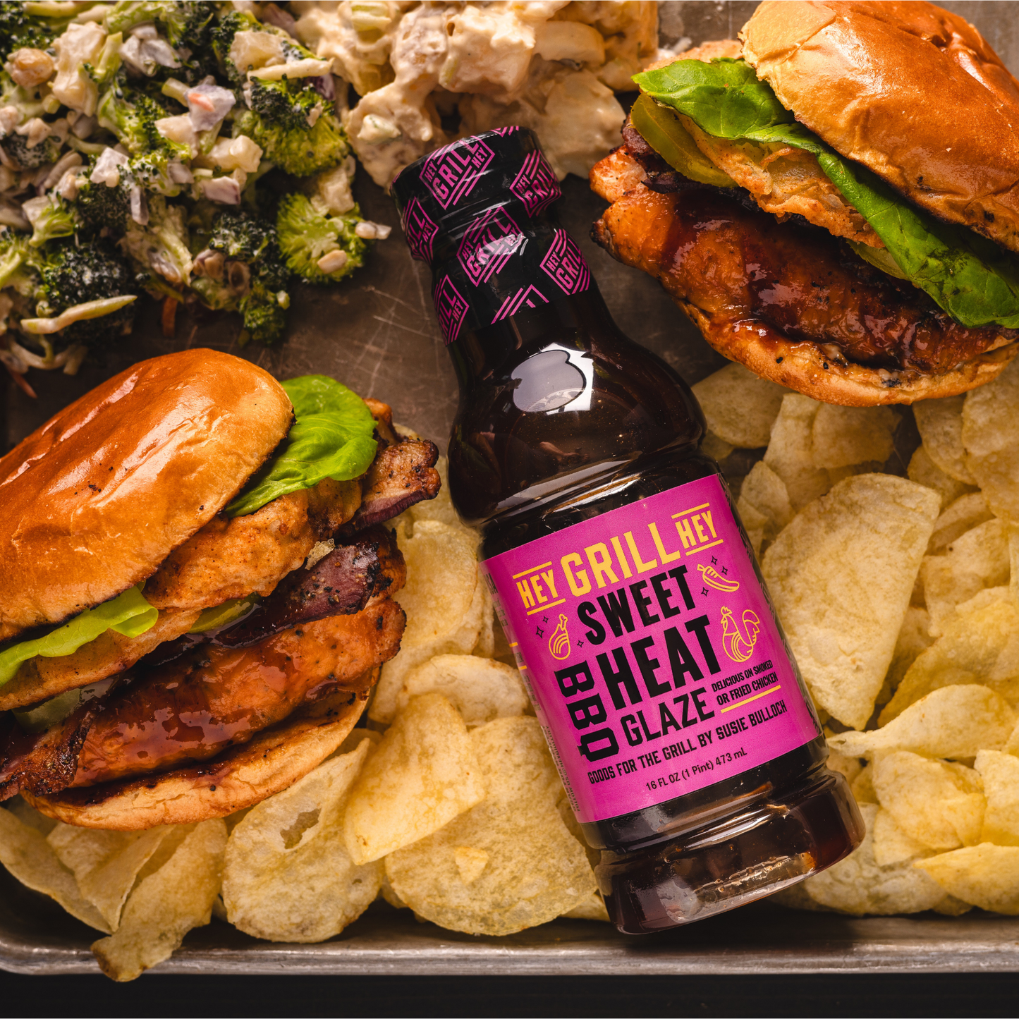 Two grilled chicken sandwiches plated with a bottle of Sweet Heat BBQ Glaze, potato chips, and a broccoli salad. 