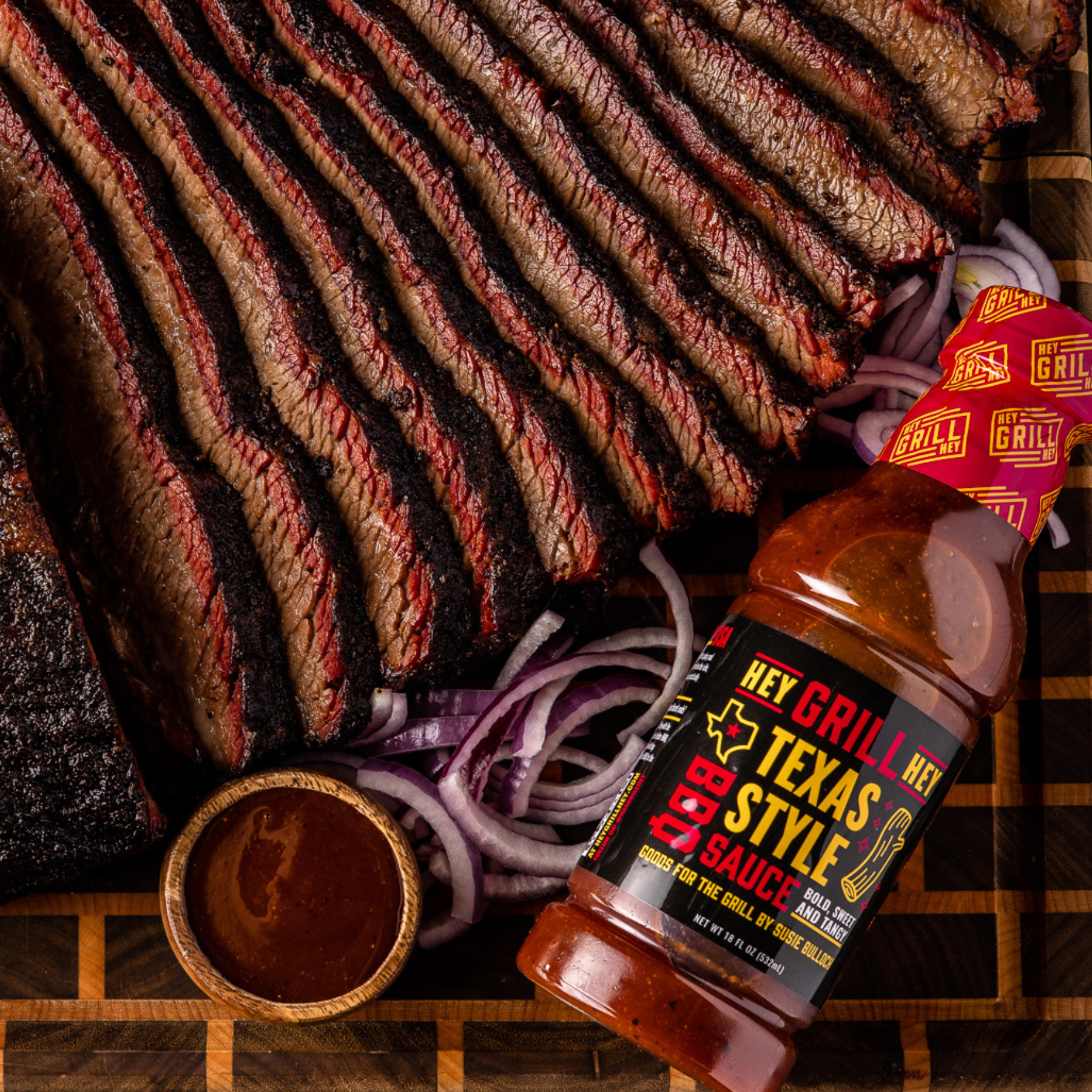 Sliced Smoked Brisket on a cutting board next to a bottle of Texas Style BBQ Sauce, garnished with onions and a small cup of BBQ sauce. 