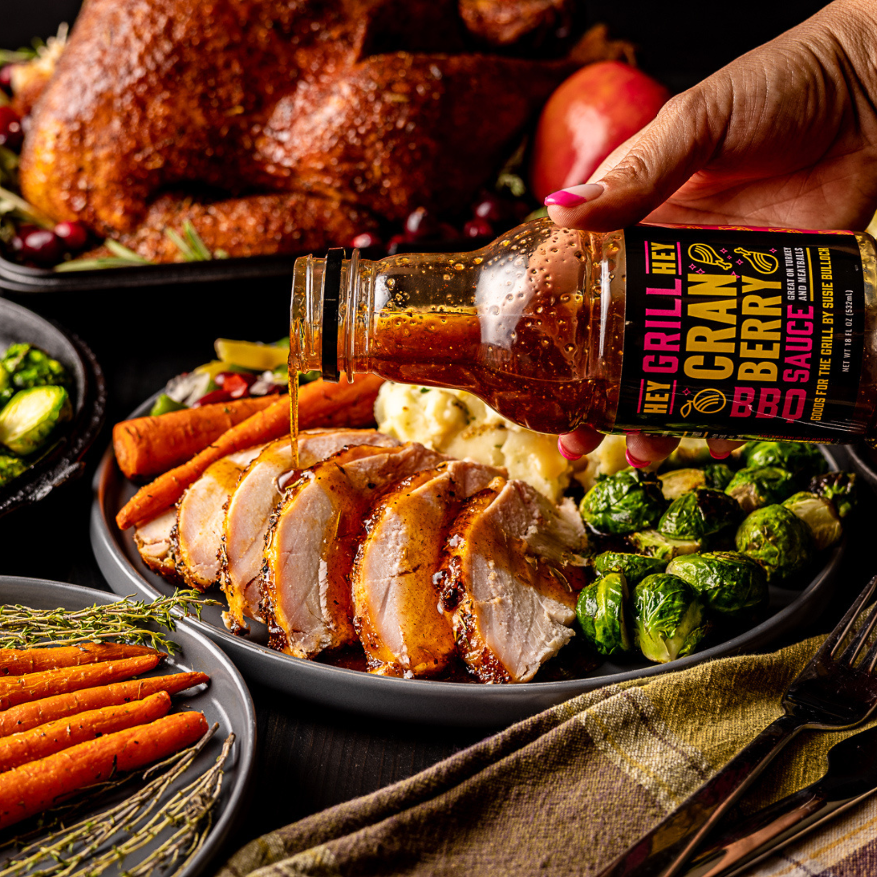 A hand holding a bottle of Cranberry Glaze pouring it over a plate with sliced turkey and other traditional sides. 