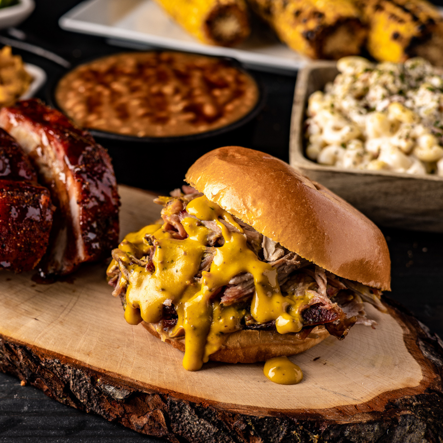 A pulled pork sandwich with Zesty Gold BBQ sauce on it sits plated on a table with other side dishes in the background. 