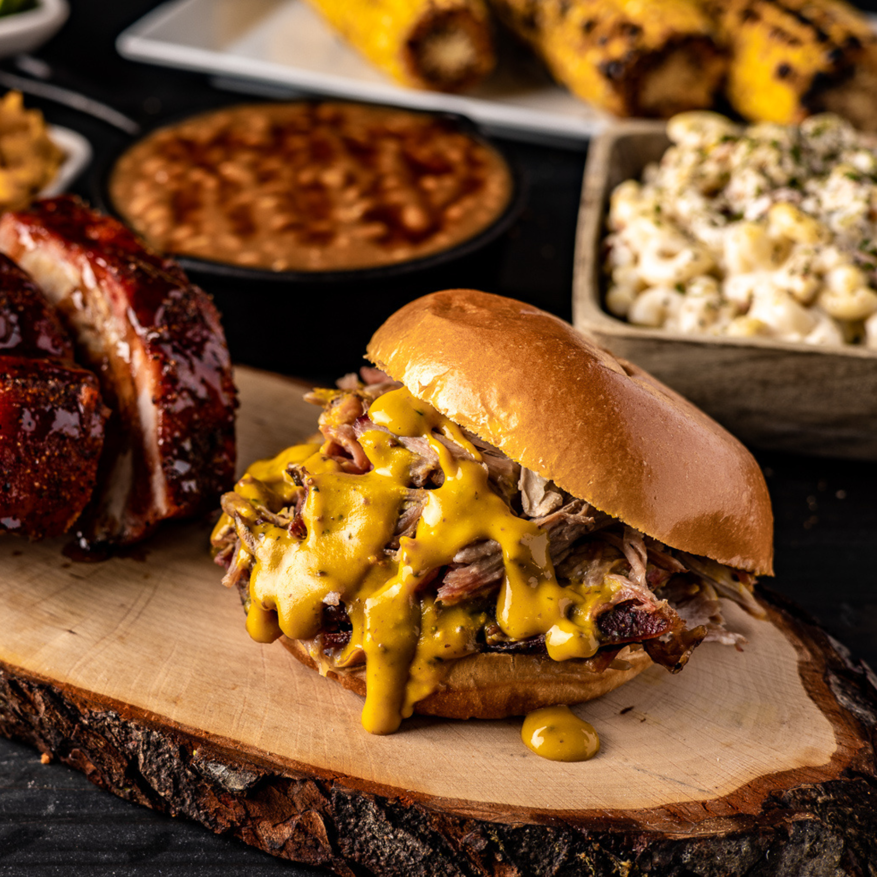 A pulled pork sandwich with Zesty Gold BBQ sauce on it sits plated on a table with other side dishes in the background. 